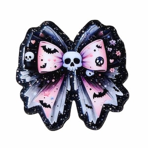 May include: A decorative bow with a skull in the center, featuring a black and pink color scheme. The bow is adorned with bats, skulls, and stars. The design has a glittery finish, adding a touch of sparkle.