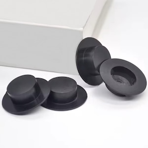 May include: Several black rubber stoppers with a hat-like design are scattered on a white surface. The stoppers have a circular base and a cylindrical top, with one stopper showing the interior. The stoppers are of varying sizes.