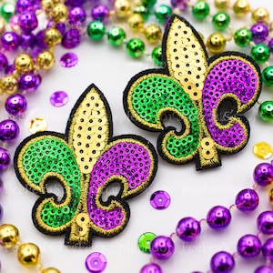 May include: Two fleur-de-lis patches featuring gold, green, and purple sequins with black embroidered edges, surrounded by strands of metallic gold, green, and purple plastic beads and loose matching sequins on a white background.