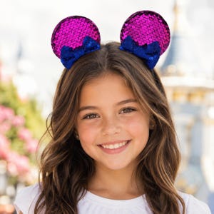 May include: A headband featuring two circular ears covered in bright magenta sequins. Each ear is accented with a small, dark blue sequined bow positioned at the base. The headband is designed to be worn on the head, providing a decorative accessory.
