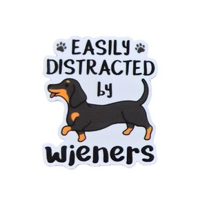 May include: A die-cut sticker featuring a black and tan dachshund illustration. The text reads EASILY DISTRACTED by WIENERS in black capital and lowercase letters. Two small black paw prints are positioned on either side of the top text line.