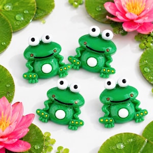 May include: Four green frog figurines with white bellies and large, white-and-black eyes. The frogs are surrounded by lily pads and pink water lilies. The figurines have a glossy finish and are detailed with yellow accents.