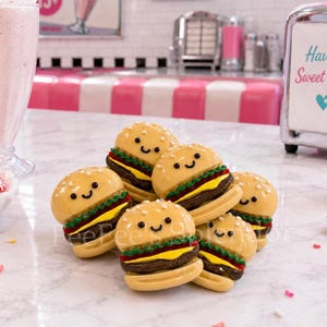May include: A cluster of six miniature burger-shaped figurines with smiling faces. The burgers have sesame seed buns, red lettuce, yellow cheese, and a brown patty. The background includes a diner setting with pink and white seating.