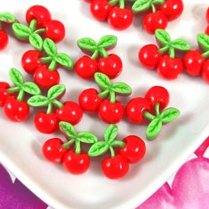 May include: A collection of small, glossy red cherry-shaped charms, each featuring two round red fruits attached to a pair of bright green leaves. These decorative items are scattered across a white surface, showcasing a smooth, shiny finish.