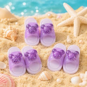May include: Several pairs of lavender and white children's sandals with glittery purple Mickey Mouse-shaped accents are arranged on a sandy beach. Seashells and a starfish are also visible in the scene.