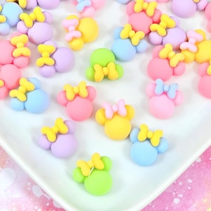 May include: A collection of colorful, pastel-colored mouse-shaped embellishments with yellow bows. The embellishments are in shades of pink, blue, yellow, green, and purple. They are arranged on a white surface.