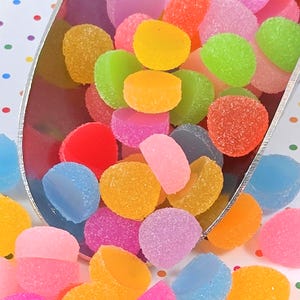 May include: A close-up shot of a variety of colorful, round gummy candies. The candies are in shades of pink, orange, yellow, green, blue, and purple. The candies have a sugary coating and are in a silver scoop.