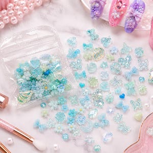 May include: A collection of light blue and clear nail art decorations, including flowers, bows, butterflies, and hearts. Some are in a clear plastic bag. The decorations are scattered on a marble surface, with a makeup brush and nail art samples in the background.