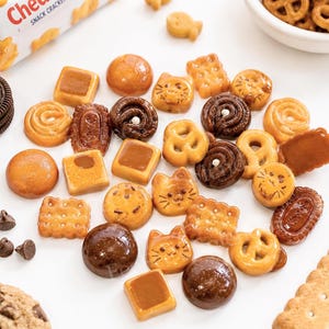 May include: A collection of miniature, realistic-looking cookie and cracker charms in various shapes including cat faces, pretzels, squares, and circles. The pieces feature shades of golden brown and dark chocolate brown with glossy, textured finishes.