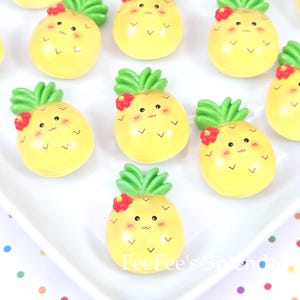 May include: A collection of yellow pineapple-shaped ornaments with green leafy tops and red flower accents. Each ornament has a smiling face and small decorative details. The ornaments are arranged on a white surface.