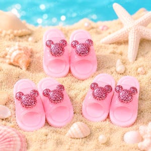 May include: Pink children's sandals with a glittery mouse head design. The sandals are arranged on a sandy surface with seashells and a starfish, suggesting a beach theme. The sandals are made of a translucent pink material.