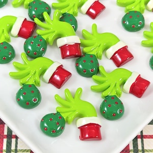 May include: A collection of small, flat-backed resin cabochons shaped like green, four-fingered hands emerging from red sleeves with white cuffs. Each hand holds a round, dark green ornament decorated with small red dots, featuring a glossy finish.