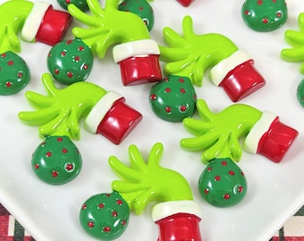 May include: A collection of green Grinch-themed ornaments and hand-shaped decorations. The ornaments are green with red dots, and the hands have red cuffs with white trim. The items are arranged on a white surface.