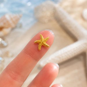 May include: A small, five-pointed golden starfish charm with a textured, granular surface. The starfish features a classic radial symmetry with tapered arms and a central disc, displaying a bright metallic yellow finish that catches the light.