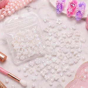 May include: A collection of iridescent nail art decorations, including bows, stars, and flowers, scattered on a surface. A clear bag filled with similar embellishments is also present. The decorations are in various sizes and shapes, perfect for nail art.