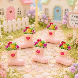 May include: Miniature pink rain boots filled with colorful flowers, arranged on a cobblestone path. Each boot has a heart-shaped detail. The scene includes miniature houses and a sign that reads "Spring Blooms This Way."