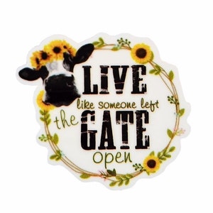 May include: A white circle with a black and white cow's head peeking out of the top. The circle is surrounded by a wreath of green leaves and yellow sunflowers. The text "LIVE like someone left the GATE open" is printed in black on the white circle.