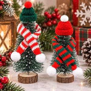 May include: Two miniature artificial Christmas trees on round wooden bases. The left tree wears a green knit hat with a red pom-pom and a red-and-white striped scarf. The right tree wears a red knit hat with a white pom-pom and a red-and-green striped scarf.