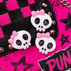 May include: Three white skull-shaped embellishments with black eye sockets and pink bows. The skulls are on a black and pink background with studs, a chain, and a torn paper with the word "PUNK".