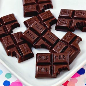 May include: Dark brown chocolate bars arranged on a white plate. Each bar is divided into four squares, with the word "MILK" embossed on each. Some bars have a bite taken out of them.