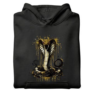 Könnte beinhalten: Schwarzer Kapuzenpullover mit goldenem Kobra-Design. Das Bild zeigt auch eine schwarze Baseballkappe, eine schwarze Jogginghose mit Kobra-Logo und mehrere schwarze Hemden. Das Kobra-Design hat goldene Akzente.