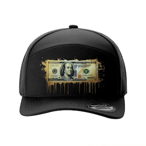 May include: Black baseball cap featuring a graphic of a $100 bill with gold paint drips. The cap has a curved brim and a snapback closure. Several other colored caps are shown in the background.