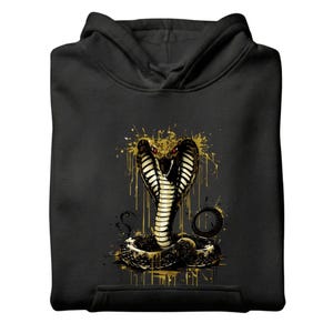 May include: A black hooded sweatshirt featuring a graphic of a cobra with red eyes and gold paint splatter effects. The design includes a black cap and black sweatpants with a matching cobra emblem. The text ALWAYS AUTHENTIC is visible.