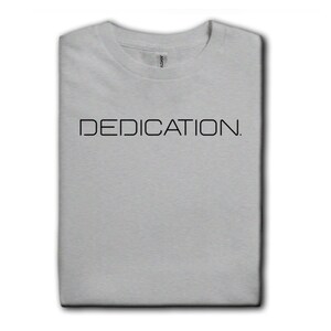 May include: A light gray short-sleeved t-shirt featuring the word DEDICATION. printed in black capital letters across the chest. The shirt is folded neatly and displayed alongside a vertical strip of various other solid color t-shirt options.