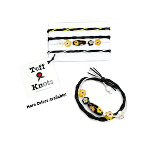 May include: Black and white braided bracelets with floral and oval beads. One bracelet is displayed separately, and others are on a white card. The tag reads "Tuff Knots" with "More Colors Available!"