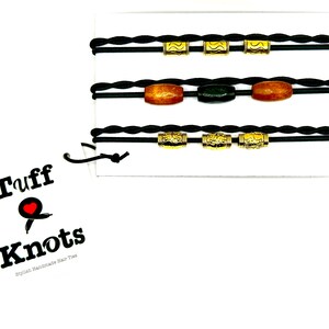 Peut inclure: Trois &eacute;lastiques &agrave; cheveux tress&eacute;s noirs avec des perles dor&eacute;es et ambr&eacute;es. Les &eacute;lastiques &agrave; cheveux sont pr&eacute;sent&eacute;s sur un fond blanc avec un logo qui dit "Tuff Knots,  Stylish Handmade Hair Ties".