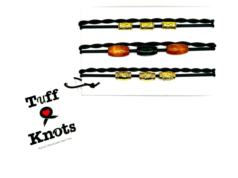 Peut inclure: Trois &eacute;lastiques &agrave; cheveux tress&eacute;s noirs avec des perles dor&eacute;es et ambr&eacute;es. Les &eacute;lastiques &agrave; cheveux sont pr&eacute;sent&eacute;s sur un fond blanc avec un logo qui dit "Tuff Knots,  Stylish Handmade Hair Ties".