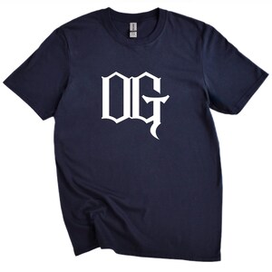 May include: Navy blue t-shirt with the white letters "OG" in a stylized font. The short-sleeved shirt is made of a soft material and is suitable for casual wear.