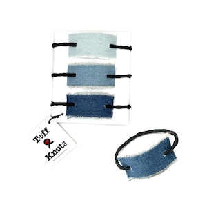 Può includere: Una scheda bianca con tre campioni di tessuto denim in diverse tonalità di blu, fissati con filo nero. Sono visibili anche una piccola etichetta in denim con un cordoncino nero e un'etichetta con il testo "Tuff & Knots".