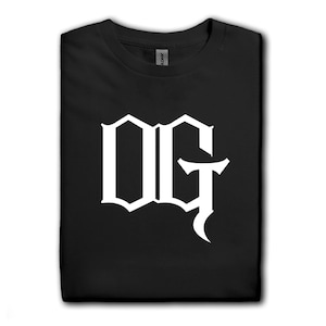 May include: Black t-shirt with a white "OG" graphic in a gothic font. The t-shirt is folded neatly, with a tag visible at the collar. Additional t-shirts in various colors are visible in the background.