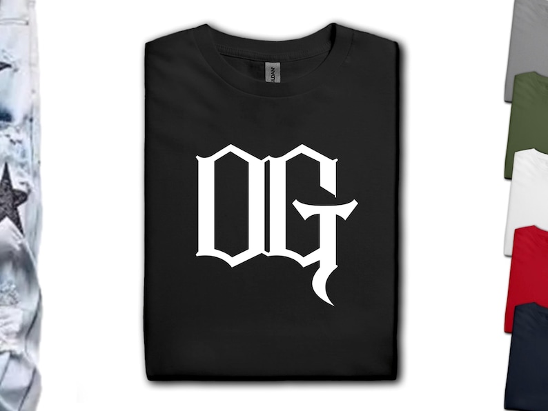 May include: Black t-shirt with a white "OG" graphic in a gothic font. The t-shirt is folded neatly, with a tag visible at the collar. Additional t-shirts in various colors are visible in the background.