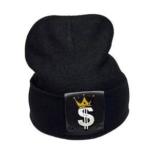 Puede incluir: Un gorro de punto negro con un parche rectangular negro en el dobladillo frontal. El parche muestra un símbolo de dólar blanco coronado por una corona amarilla. El texto "Undeniably Uncommon..." aparece a la izquierda y "ALWAYS AUTHENTIC" a la derecha.