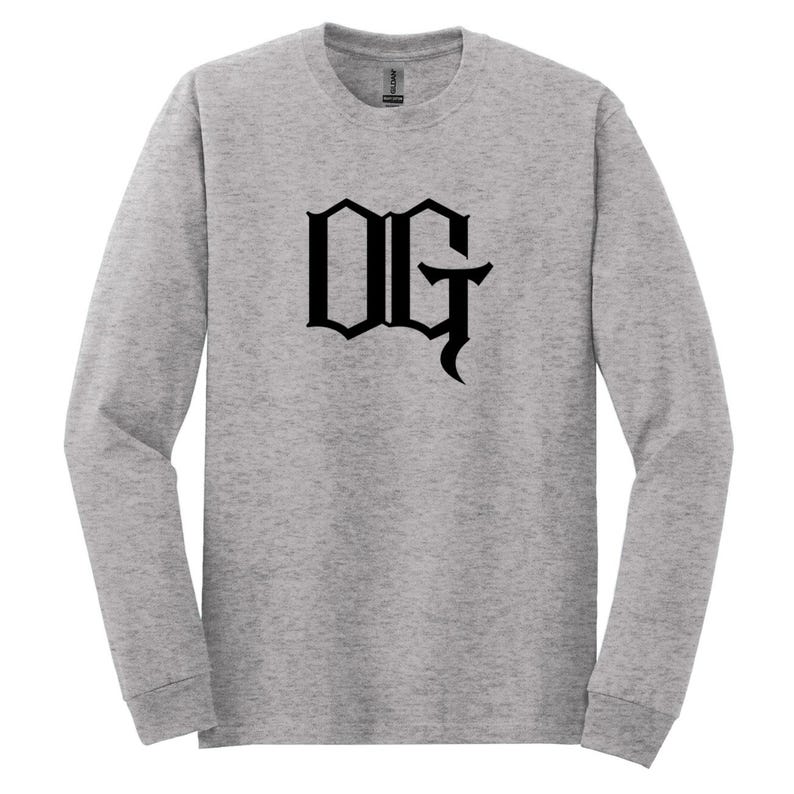 May include: A light gray long-sleeve shirt with the letters "OG" in a bold, black, gothic font centered on the front. The shirt has a crew neck and long sleeves.