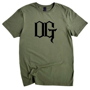 May include: Olive green t-shirt with the black letters "OG" in a gothic font. The short-sleeved shirt is made of a soft material and is displayed on a white background. The shirt is a casual wear item.