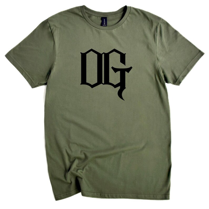 May include: Olive green t-shirt with the black letters "OG" in a gothic font. The short-sleeved shirt is made of a soft material and is displayed on a white background. The shirt is a casual wear item.