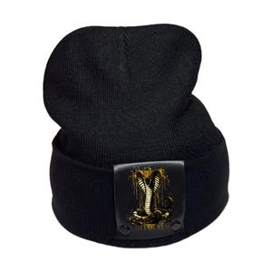 May include: A black knit beanie featuring a rectangular leather-like patch on the front. The patch displays a graphic of a cobra snake in shades of gold and black with a dripping paint effect. The text "Undeniably Uncommon..." and "ALWAYS AUTHENTIC" appear.
