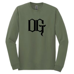 May include: Olive green long-sleeve shirt with the black letters "OG" in a gothic font. The shirt is made of a soft material and has a crew neck. The shirt is a comfortable and casual style.