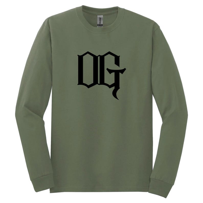 May include: Olive green long-sleeve shirt with the black letters "OG" in a gothic font. The shirt is made of a soft material and has a crew neck. The shirt is a comfortable and casual style.