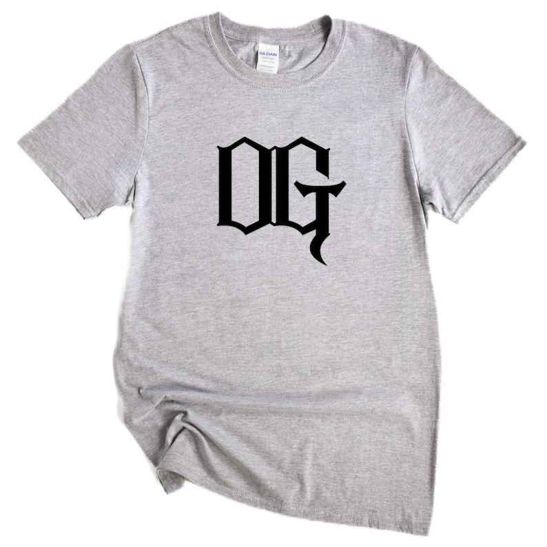 May include: A light gray t-shirt with the black letters "OG" in a gothic font. The short-sleeved shirt is made of a soft material and is suitable for casual wear. The shirt is displayed on a white background.