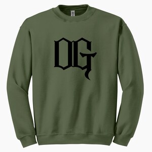 May include: Olive green sweatshirt with the black letters "OG" in a gothic font across the chest. The sweatshirt has a crew neck and long sleeves. The fabric appears to be a soft, comfortable material.