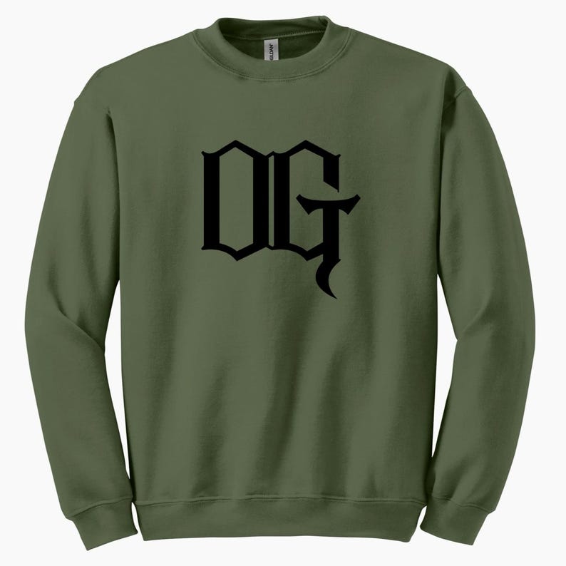 May include: Olive green sweatshirt with the black letters "OG" in a gothic font across the chest. The sweatshirt has a crew neck and long sleeves. The fabric appears to be a soft, comfortable material.