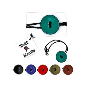 Könnte beinhalten: Handgemachte Haargummis mit teal-farbenen Knöpfen und dem Label "Tuff & Knots". Das Bild zeigt auch Haargummis mit schwarzen, blauen, roten, olivgrünen und braunen Knöpfen, alle mit schwarzem Gummiband.
