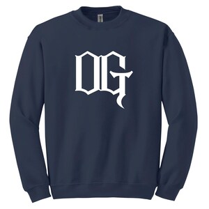 May include: Navy blue sweatshirt with the white letters "OG" in a gothic font. The crew neck sweatshirt is a solid color and has long sleeves. The letters are centered on the front of the shirt.