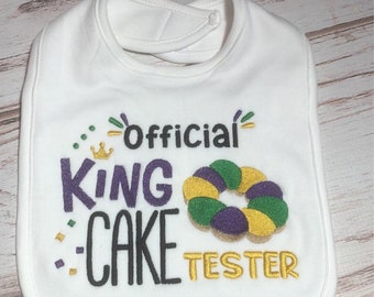 May include: White baby bib with the embroidered text "Official King Cake Tester" in purple, black, and gold. A gold crown and a king cake graphic are also embroidered on the bib.