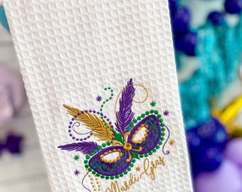 May include: White kitchen towel with an embroidered purple and gold Mardi Gras mask and the text "Mardi Gras".