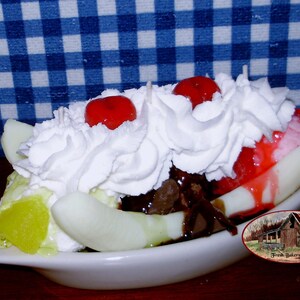 May include: A banana split candle in a white dish. The candle features a banana base, topped with whipped cream, red cherries, chocolate sauce, and green and red ice cream. The candle is a dessert-themed item.